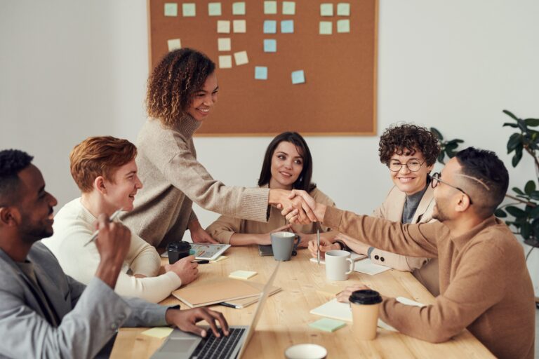 Group of 6 university students, female and male, at a table. Two are shaking hands across the table.